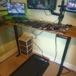 An elevated standing desk holds a laptop and various devices with a walking pad underneath